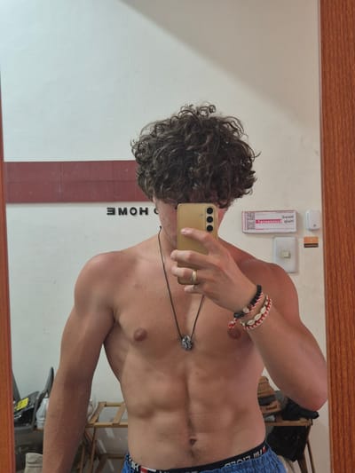 A tanned shirtless man with curly hair is taking a mirror selfie with a gold-colored smartphone. He is wearing blue shorts with a red, white, and blue waistband, a black necklace with a pendant, and several bracelets on his left wrist. A gold ring is visible on his left hand. The word "HOME" is visible on the wall behind him. A light switch, outlet, and a small white box are on the wall above him. There is a chair and some other objects in the background. A small red and white "Need Help" card with a QR code is visible on the wall. The location is Pasay, Philippines.