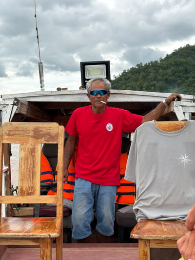 In Xanghay, Laos, a man stands on a boat, smoking a cigarette and wearing blue sunglasses, a red t-shirt, and denim shorts. He is leaning against the boat's structure, with his left arm resting on it. His right arm is extended, with a gray t-shirt hanging on a wooden chair in front of him. The background shows a cloudy sky and a lush green hillside. Orange life vests are visible behind the man, suggesting the boat is used for transport or a tour. The overall scene appears to be a casual, outdoor setting on the water.