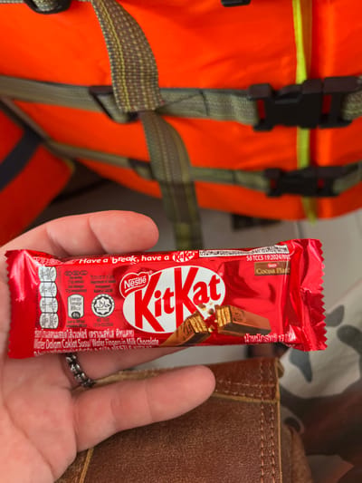 The image captures a close-up, downward-angled view of a light-toned left hand holding a red-packaged Nestlé Kit Kat chocolate bar. A silver-toned ring is worn on the ring finger. The Kit Kat wrapper features the "Nestlé Kit Kat" logo, the slogan "Have a break, have a Kit Kat," an illustration of chocolate fingers, and nutritional details including a Halal certification and text in English and Thai, specifying "17 กรัม" (17 grams) for weight. In the blurred background, an orange life vest with dark green straps and black plastic buckles is visible, occupying the upper portion of the frame. Below the hand, a section of a brown, stitched material, possibly leather or synthetic, is present. The setting, characterized by the life vest, indicates a marine environment, likely on a boat or dock in Sihanoukville, Cambodia.