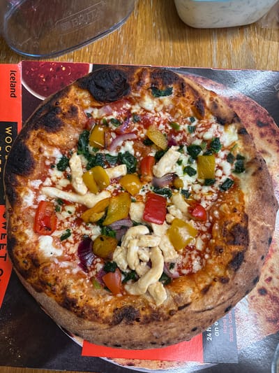 The image shows a pizza on top of a cardboard pizza box. The pizza has a charred crust and is topped with tomato sauce, mozzarella cheese, red onions, yellow bell peppers, red bell peppers, spinach, and pieces of chicken. The pizza box has branding and text visible, including the word "Iceland" and parts of words such as "WOOD" and "SWEET". There is also a Brita water filter pitcher and a bottle of ranch dressing on the wooden table next to the pizza and pizza box.
The photo was taken on a table, and the pizza is sitting on top of its original cardboard box. There are no people visible in the image. The location in the city of Oswestry, United Kingdom, is not mentioned in the image or description.