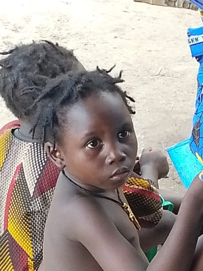 This media file shows a close-up of two young children in what appears to be an outdoor setting in Otulu, Nigeria. The child in the foreground is facing the camera, looking directly at it with a serious expression. This child has dark skin, dreadlocks, and is wearing a necklace with a gold-colored pendant. They are also wearing a colorful garment with a geometric pattern in red, yellow, black, and white. Part of another child is visible behind the first, also with dreadlocks and wearing a similar patterned garment. The background is a sandy or dusty ground. To the right, a portion of a blue object is visible, possibly a piece of clothing or an accessory, with some text that is partially legible. The visible text appears to be "SEK K" in white letters on a blue background, though it is cut off and difficult to decipher fully. The lighting suggests it might be daytime, and the overall impression is of a candid moment captured outdoors.