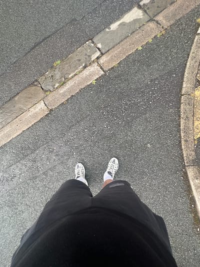 The image shows a person's legs and feet standing on a wet asphalt road in Wrexham, United Kingdom. The person is wearing black shorts and white and black athletic shoes with white socks. The road has a concrete curb on the right side, which is partially yellow. A concrete kerb runs along the left side of the road. There are some small weeds growing between the kerbstones. The road surface appears to be textured with small stones. The lighting suggests it is daytime, possibly overcast or after rain due to the wetness. The perspective is from a high angle, looking down at the person's lower body and the immediate surroundings.