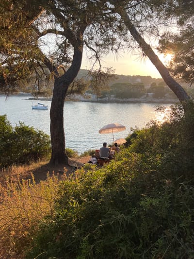 A tranquil scene shows a body of water framed by trees. A group sits under a parasol at the edge of the water, enjoying a moment by the lake. A small sailboat is visible in the water. The sun reflects on the surface, adding to the serene atmosphere. The foliage is lush on one side.