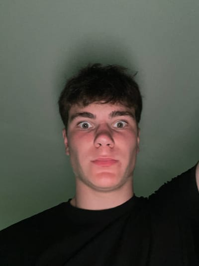 The image is a close-up, front-facing selfie of a young male with dark, short hair and wide, surprised eyes. He is wearing a black t-shirt. The background is a plain, muted green wall. The lighting is from above and appears to be artificial, casting a shadow on the wall behind him. There are no other discernible objects or people in the image. The location is not specified or discernible from the image itself.