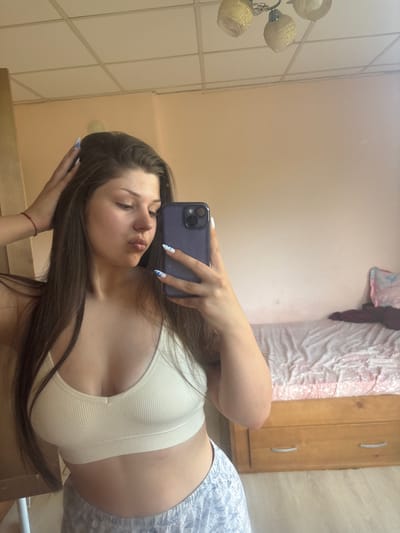 The image shows a young woman taking a selfie in a dimly lit room. She is wearing a white ribbed sports bra and patterned pajama bottoms. Her long, dark hair is down, and she is leaning her head to the side, with her eyes closed and lips pursed in a playful expression. Her fingernails are painted with a white and blue design. She is holding a dark purple smartphone, capturing her reflection in a large mirror that takes up much of the left side of the frame.
The background is a bedroom. To the left of the woman, in the mirror's reflection, is a ceiling light fixture with three glass lampshades and a metal base. Further into the reflection, we see a tiled wall. To the right of the woman, outside the mirror's reflection, is a wooden wardrobe with two doors and simple silver handles. Next to the wardrobe, on the floor, is a rumpled pink and white patterned duvet or blanket. The overall impression is of a casual, domestic setting, likely a bedroom. The lighting suggests it could be daytime, but the indoor setting and the use of a phone to take the picture don't provide definitive clues about the time of day or weather. The woman's expression and pose suggest a relaxed and possibly playful mood. There is no visible text in the image. The location context is Prodanovtsi, Bulgaria.