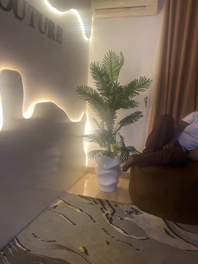 The image captures an indoor scene, likely a modern living room or lounge area, during the evening or night, as indicated by the prominent use of artificial lighting. The location is Dushepe, Nigeria.
A person is visible, seated on a dark brown or olive-green sofa or plush chair, facing away from the camera. Only their lower torso and legs are visible; they are wearing a white t-shirt and dark brown trousers. Their bare feet are relaxed on the floor, and their toenails have white polish.
The room features a contemporary design. A large, artificial palm-like plant with vibrant green fronds stands in a stylish white, multi-tiered vase on the floor, positioned near the seated person. The walls are a light, neutral color, possibly white or off-white. Unique, organic-shaped wall panels are a dominant feature, integrated with hidden LED strip lighting that casts a warm, soft glow, highlighting their contours. Part of a word is visible on one of these illuminated panels, spelling "OUTURE," suggesting "COUTURE."
An air conditioning unit is mounted on the wall in the upper left section of the image. Dark brown curtains or blinds are drawn across the upper part of the wall, possibly covering a window. The floor is partially covered by a modern rug featuring abstract, curvilinear patterns in shades of grey, black, and hints of light brown or gold. The overall ambiance is cozy and sophisticated, emphasized by the indirect lighting and natural elements like the plant.