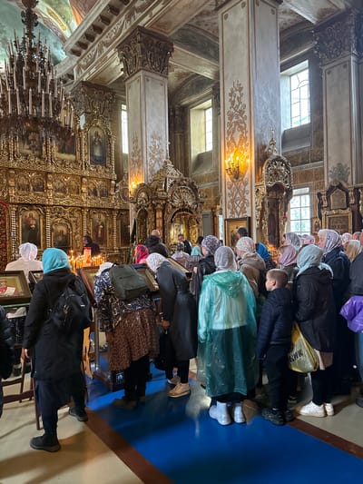 The image depicts the interior of an ornate Orthodox church, likely within the city of Раифа, Russia. A group of people, predominantly women, are gathered in front of a large, gilded iconostasis adorned with multiple religious icons and lit candles. Many are wearing headscarves, and some are kneeling or bowing, engaging in prayer or veneration. The church architecture is richly decorated with columns featuring intricate carvings and gold accents, and the ceiling is painted with frescoes. Sunlight streams in through tall, arched windows. The floor is covered in a vibrant blue carpet. The overall atmosphere suggests a religious service or a period of devotion.