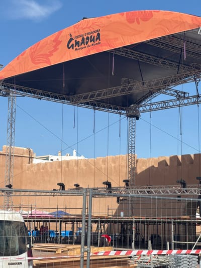 The image shows an outdoor stage under construction in Essaouira, Morocco. The stage features a large orange banner with the text "FESTIVAL D'ESSAOUIRA Gnaoua Musiques du Monde." The stage structure is made of metal trusses and scaffolding, with a dark fabric covering the top. In the background, there is a characteristic terracotta-colored wall, typical of Essaouira's historic medina and ramparts. Beyond the wall, white buildings with flat roofs are visible, also consistent with local architecture. The sky is clear and blue, with a small airplane visible in the distance. In the foreground, construction fencing is in place, with stacks of metal scaffolding and wooden pallets. A white van is parked to the left, with "DOKKER" visible on its rear. Several people are present, some working on the construction and others appearing to be seated behind the fencing, possibly spectators or staff. The overall scene suggests preparations for an outdoor event.