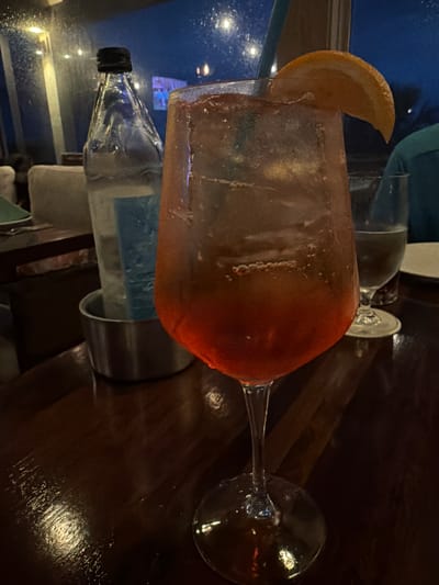 A close-up, eye-level shot captures a glass of Aperol Spritz on a dark wooden table. The drink is in a large wine glass and filled with ice. A thin blue straw extends from the glass, and an orange slice is perched on the rim. In the background, a bottle of sparkling water sits in a silver bucket. The scene appears to be set in a restaurant or bar on Hilton Head Island, South Carolina, given the ambiance suggested by the dimly lit environment and the style of the establishment. The lighting suggests it is evening or night.