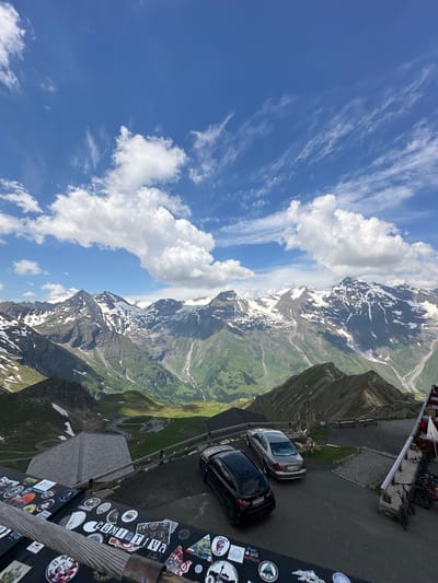 The image depicts a scenic mountain landscape in Fusch an der Großglocknerstraße, Austria, with a parking area in the foreground. The sky is a vibrant blue with scattered white clouds. The mountains in the background are majestic, with snow-capped peaks and green valleys. A winding road is visible in the distance, snaking through the mountains.

In the foreground, there is a black SUV and a silver sedan parked on an asphalt surface. A wooden railing separates the parking area from a steep drop-off. Several bicycles are parked to the right of the frame, leaning against a building. The surface in the immediate foreground is covered with numerous stickers, suggesting it might be a popular stop for tourists. A building with a sloped, grey roof is visible on the left side of the parking area. The overall impression is one of a popular viewpoint offering breathtaking mountain vistas.