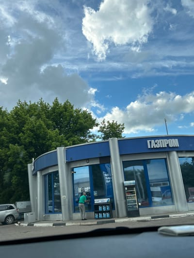 The image shows a Gazprom gas station in Novobeisugskaya, Russia. The gas station has a blue and silver facade with the Gazprom logo displayed prominently. A man dressed in a green shirt and light-colored pants is standing at an automated teller machine. The sky above shows a mix of blue sky and clouds. There's a tree positioned between the sky and the gas station. Part of a silver car is visible in the lower right corner. The image was taken from inside another vehicle.