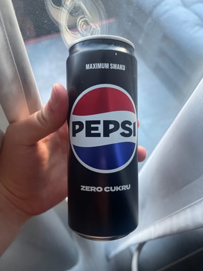A hand holds a black Pepsi Zero Sugar can. The can has the Pepsi logo, which is a red, white, and blue circle. Above the logo, the text "MAXIMUM SMAKU" is printed in white. Below the logo, the text "ZERO CUKRU" is printed in white. The can is held in front of a blurred background that appears to be the interior of a vehicle, with white fabric visible on the right and a window on the left. The lighting suggests it is daytime.