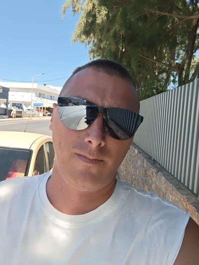 The image is a bright, sunlit selfie taken outdoors of a man in Gouves Municipal Unit, Greece. The man, who appears to be in his late 20s or 30s, is positioned centrally in the frame, looking directly at the camera with a neutral expression. He has a short, dark buzz cut and is wearing large, mirrored sunglasses that reflect the surroundings. He is dressed in a plain white sleeveless t-shirt or tank top.

To the left, partially visible behind his shoulder and arm, is the rear section of a light-colored car, possibly yellow or beige, with a red or maroon interior. In the background, across the street, white buildings with various windows and awnings are visible under a clear blue sky, along with a street lamp. The mirrored sunglasses clearly reflect the paved road, a distant person in a light outfit, and a small phone or camera held by the man, capturing the selfie.

On the right side of the frame, behind the man, is a grey and white vertically striped fence, built upon a textured stone wall. Lush green foliage from trees, possibly conifers, extends into the upper right and top center portions of the image. The bright light and clear blue sky indicate it is a sunny daytime scene, likely mid-day. Visible Greek text on a sign above one of the buildings across the street reads "πωληση πωληση," which translates to "sale sale" or "for sale for sale." The scene depicts a typical street environment in a warm climate.