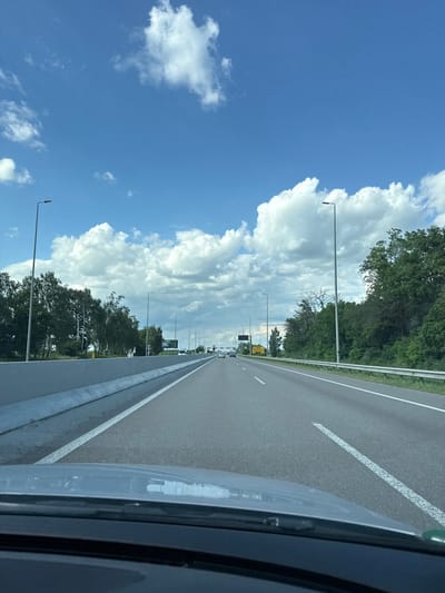 The image depicts a view from inside a vehicle traveling on a highway. The highway has two lanes in the direction of travel, marked by white lines. To the left of the highway is a grassy shoulder bordered by a metal guardrail. Beyond the guardrail, a bright blue sky is filled with large, fluffy white clouds. Tall trees line the left side of the highway, their green foliage contrasting with the sky. Lampposts are visible along the roadside. In the distance, there are more trees and some structures. The right side of the image shows the side of the vehicle, including the side mirror and a portion of the car's body. The scene suggests a daytime drive on a clear, possibly warm day, with a mix of natural scenery and infrastructure. The orientation of the image is rotated 90 degrees counter-clockwise. There is no visible text in the image.