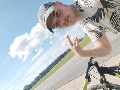 A young man wearing a grey baseball cap and a t-shirt with a black and white palm leaf pattern is taking a selfie. He is on a bicycle, holding up two fingers in a peace sign. The background shows a blue sky with scattered clouds, a green field, and a line of trees. The sun is shining, suggesting it is daytime. The man appears to be outdoors, possibly on a road or path, given the visible asphalt. The image is taken from a low angle, looking up at the man and the sky. The bicycle is partially visible in the foreground. The man's t-shirt has a pattern of black leaves on a white background. The baseball cap has white text or a logo on it, but it is not legible. The overall mood seems casual and cheerful.