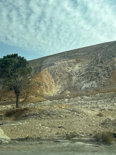 The image displays a rugged, arid landscape situated within the city of Ma'raba, Syria. The sky occupies the upper portion, featuring a light blue hue with numerous scattered white cirrocumulus clouds. The dominant feature is a steep, rocky hillside or mountain face that extends across the upper two-thirds of the frame. The upper slope appears dark grey, likely indicating sparse vegetation or shadowed rock. Below this, the hillside presents distinct geological features: a prominent section of light brown-tan rock on the left transitions into exposed white, light grey, and dark grey rock formations with visible stratifications and erosion patterns. Small, faint, dark rectangular openings, possibly caves or structures, are visible within the rock face, and a subtle, light-colored building-like structure is faintly discernible on the lower-mid brown section of the hill.

The lower third of the image constitutes a disturbed foreground composed of loose soil, gravel, and various sizes of rock debris, forming uneven mounds and a generally uncultivated surface. A single, mature conifer tree with dense green foliage stands on a raised earthen mound in the lower-left quadrant. The very bottom of the frame reveals a horizontal concrete edge or barrier, suggesting the immediate proximity of a paved surface or road. No individuals or active interactions are present in the scene.