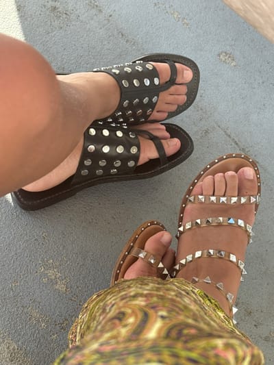 This image, captured in Ribamontán al Mar, Spain, presents a close-up, overhead view of two individuals' feet resting on a textured, light grey concrete or paved surface.
The subject on the left wears black, stud-adorned flat sandals. These sandals feature a wide forefoot strap with multiple rows of circular, silver-toned studs and a thin toe-post. A portion of the individual's lower leg, exhibiting a fair to light-tanned skin tone, is visible above the left foot.
The subject on the right wears light brown or clear flat sandals with multiple transparent straps, each embedded with pyramidal, silver-toned studs. This individual's foot and ankle are visible, partially obscured by a garment displaying a vibrant, multi-colored swirling pattern, likely a skirt or dress.
Both pairs of feet are positioned side-by-side, suggesting two individuals standing or sitting in close proximity. The concrete surface beneath them shows minor variations in texture and color.