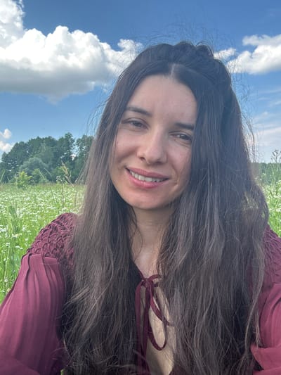 A selfie is taken by a woman with long, dark hair, wearing a maroon shirt, in a field of white flowers under a blue sky with clouds. The background consists of green trees. The location is Vyshevychi, Ukraine.