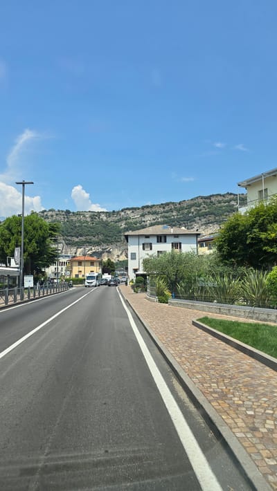 This is a photograph taken from a vehicle, likely moving along a road. The scene is oriented vertically, suggesting the original photo was taken sideways. The road surface is asphalt with a white line marking. To the left of the road, there is a low wall constructed from small, multi-colored stones, topped with a strip of green grass. Further left, a steep, rocky mountainside covered in vegetation rises sharply. Several buildings are nestled at the base of the mountain, ranging from multi-story white structures to smaller, colored houses. Trees, some with dense green foliage, are scattered throughout the scene. The sky is a bright, clear blue with a few wispy white clouds. The overall impression is of a sunny day in a mountainous, possibly coastal, region. Text is visible on a sign near a lamppost at the bottom left of the image, though it is illegible. Another sign is visible on the white building in the middle left, showing a circular logo with stylized letters, also illegible. The lighting suggests it is daytime, likely mid-morning or afternoon. The presence of vehicles on the road indicates it is a public thoroughfare. The setting is consistent with a town like Torbole, Italy, known for its lakeside location and proximity to mountains. There are no people clearly visible in the frame.