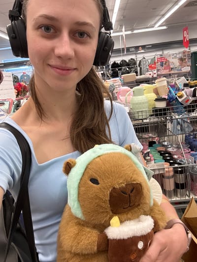 A young woman wearing headphones and a light blue top is posing for a selfie in a store. She is holding a stuffed capybara with a light blue bandana and a brown cup with white trim. In the background, there are shelves stocked with various items, including water bottles and bags, and signage indicating discounts. The location appears to be a retail store in Erlenbach am Main, Germany, as suggested by the German text on some of the signs and the overall appearance of a typical German department or discount store.