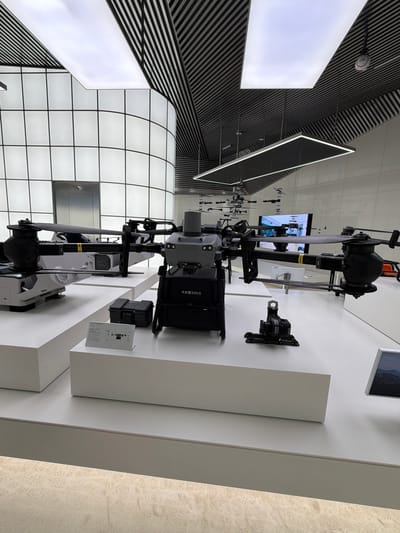 This media file captures an indoor exhibition of advanced drone technology, likely in a modern commercial or event space. The setting features a sleek, minimalist design with white, angular display platforms against a backdrop of black and white striped walls and a grid-patterned illuminated ceiling.

The primary objects on display are several large, professional-grade drones. One prominent drone, partially disassembled, is situated on a white pedestal, showcasing its intricate mechanical components, including rotors and a central body unit. To its right, a smaller, black drone is mounted on a wall bracket. Another drone, larger and more robust, is positioned behind the first, with visible text on its casing that appears to be a payload capacity indicator, possibly reading "可负重30KG" which translates to "Can carry a load of 30KG." A display card with a diagram of a drone and text is placed near one of the exhibits.

The lighting is bright and even, suggesting an indoor environment during daytime, though the exact time is not discernible. The overall mood is professional and technologically advanced. The image is oriented vertically, with the subjects appearing in this orientation. The location context is identified as Nanshan District, China, suggesting a sophisticated technological exhibition likely within this area known for its innovation. There are no people visible in the frame, emphasizing the focus on the technology itself.