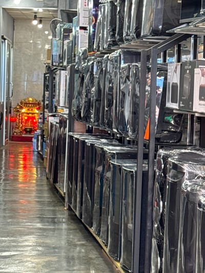 The image captures an aisle within a retail store in Ranong, Thailand, likely a electronics or appliance shop, judging by the rows of packaged goods on display. The flooring is a polished, reflective surface, likely concrete or tile, showing streaks and reflections of the overhead lighting and the items on display. To the left of the aisle, a brightly lit, ornate shrine is visible, adorned with red lanterns and gold accents, suggesting a cultural or religious element within the commercial space. The shelves are filled with various products, many of which are encased in plastic packaging. The overall impression is one of commercial organization with a touch of local cultural decoration.