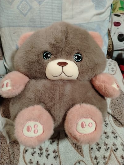 A plush brown teddy bear with pink accents sits on a patterned surface. The bear has large, round blue eyes with black pupils and light blue rims. Its muzzle is light beige, with a brown embroidered nose and a stitched mouth. The bear's ears are also pink and appear to be made of a softer, fluffier material than its body. Its paws are also pink, with white oval pads on the bottom of each, featuring embroidered pink toe beans. The bear is positioned centrally in the frame, facing forward. The background consists of pillows or blankets with a light blue and white diamond pattern, and other indistinctly patterned fabrics. The scene appears to be indoors. The image does not contain information that would allow for the determination of its location in Жернов ауылдық округі, Kazakhstan.