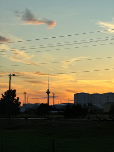 The image captures a dramatic sunset over Liivamäe, Estonia, with an industrial landscape silhouetted against the vibrant sky. In the foreground, a dark, grassy field extends towards a line of dark trees and what appears to be a fence or barrier. Overhead, several power lines cut across the frame, adding a geometric element to the scene.

Dominating the middle ground are industrial structures. To the left, a tall transmission tower with multiple antennas stands prominently, accompanied by a street lamp. Further to the right, a striking television tower with a distinctive open-lattice design rises towards the sky, also equipped with antennas. Behind these towers, the roofs of various industrial buildings are visible.

The background is characterized by a row of large, cylindrical storage tanks, characteristic of an industrial facility. These tanks are topped with numerous antennas, hinting at their function in communication or energy infrastructure. A smaller, yellow building is partially visible to the far right.

The sky is a spectacle of color, transitioning from a pale blue at the top to a brilliant orange and yellow where the sun is setting. Wispy clouds, tinged with pink and orange, add texture and depth to the celestial canvas. The overall impression is one of a vast, open sky meeting a functional, man-made environment, all bathed in the warm, fading light of dusk. No people are visible in the frame.