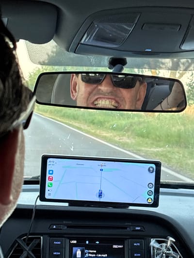 This media file depicts the interior of a vehicle, likely a car, seen from the perspective of a passenger or rear-seat occupant. The primary focus is on the dashboard area, including a rearview mirror, a mounted smartphone displaying a navigation app, and the car's infotainment system.

In the rearview mirror, a man wearing sunglasses is visible, smiling widely with his teeth bared. The reflection shows his face and part of his chest. The background reflected in the mirror appears to be an outdoor scene with green foliage and a road, suggesting the vehicle is in motion.

On the dashboard, a smartphone is mounted and shows a map interface with a route highlighted. The screen displays the time as "20:12" and indicates "5G" connectivity. Various app icons are visible on the phone screen, including what looks like a shopping cart, a person icon, and a phone icon. The number "76" is prominently displayed in a circle at the top of the navigation app, possibly indicating a speed limit or route number.

To the right of the smartphone is the car's infotainment system. Buttons labeled "DISP," "MUTE," and "SCAN" are visible, along with a slot for a USB drive and buttons for "RADIO" and "MEDIA." Text on the screen reads "[FILE] 0007" and "Home Roya - Lie.mp3," indicating music playback is in progress. The overall setting is the interior of a modern vehicle during daylight hours, as suggested by the reflections and the brightness of the outdoor scenery. The presence of the navigation app and the driver's expression imply the vehicle is on a journey. The location context provided indicates the scene is set in Stefanovo, Bulgaria.