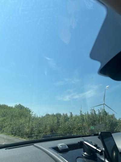 The image shows a view from inside a vehicle in Cross of The Wood, Ireland. The foreground is dominated by the dashboard of a car, including a phone mount and a phone. The windshield reflects the sky and a portion of the car's interior. Beyond the windshield, a landscape is visible under a clear blue sky with some wispy clouds. A dense line of green foliage forms a barrier across the middle ground. To the right of the foliage, a wind turbine stands tall, with its blades extended. A road with a yellow line is visible on the left side of the image, running alongside the foliage.