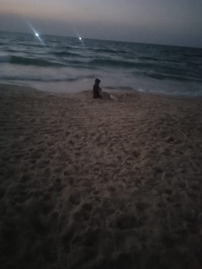 This image depicts a coastal scene at dusk or night in Al-Mawasi, Palestinian Territories. A single individual, appearing to be a young child, is seated in the midground on a sandy beach, facing the ocean. The child is dressed in dark clothing and is positioned near the waterline, engaged in an activity involving the sand directly in front of them, possibly building or playing. The foreground consists of dark, textured beach sand. In the background, the sea exhibits gentle waves breaking on the shore. Two prominent bright lights are visible on the distant horizon over the dark ocean, with a third, fainter light to the left, likely emanating from vessels or coastal structures. The sky above is dark, consistent with twilight or nighttime conditions, and the entire scene is dimly lit.