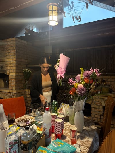 The image depicts an outdoor dining area with a brick oven and a wooden roof. A woman is standing behind a table laden with food and drinks, holding a bouquet of flowers. The table is covered with a lace tablecloth and includes various items such as juice boxes, plastic cups, a bottle of soda, and a vase with a large floral arrangement. The scene appears to be set at dusk, with artificial lights illuminating the area. The overall impression is one of a casual gathering or celebration.