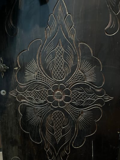 This image features a close-up view of an intricately carved black wooden surface, likely part of a chest or furniture piece. The carving is ornate and symmetrical, dominated by a floral motif in the center. This central element appears to be a stylized flower with layered petals, surrounded by elaborate scrolling foliage and fan-shaped patterns filled with cross-hatching. The carving is detailed, with fine lines creating texture and depth within the design.
The surface itself is dark, with a glossy finish that reflects light in subtle ways, highlighting scratches and imperfections that suggest age and use. The overall impression is one of traditional craftsmanship and decorative artistry. There are no people or any indication of an ongoing event. The lighting is somewhat diffused, suggesting an indoor setting or an overcast day, and no specific time of day is discernible. The location context of Bandarbireuen, Indonesia, hints at the cultural origin of such decorative wood carving, often found in traditional Indonesian furniture and art. No visible text is present in the image. A small keyhole is visible towards the bottom right of the frame, suggesting this is part of a functional item like a chest or cabinet.