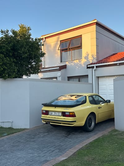 A yellow Porsche 944 is parked in the driveway of a house in Blouberg, South Africa. The house is two stories tall, with a white exterior and a brown roof. There is a window on the second floor and a garage door to the right of the car. The car's license plate reads "CAA 276-223." A tree is on the left side of the house. The driveway is paved with gray bricks. A strip of green grass is next to the driveway. The sky is blue.