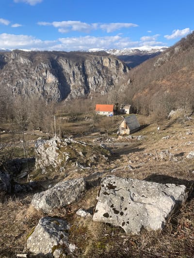 The image displays a mountainous rural landscape in Mokro, Montenegro, under a clear blue sky with scattered white clouds. The foreground features a dry, rocky incline populated by numerous large, light-grey boulders, some with moss patches, interspersed with sparse brown vegetation and dry grass. In the midground, a cluster of three structures is visible on a less steep slope. The primary structure is a single-story building with light-colored walls and an orange-tiled gable roof; a blue tarp is present adjacent to its base. Beside it, to the right, is a smaller, weathered structure with a dark, possibly wooden, gable roof. Below these, a third, smaller wooden building with a steeply pitched dark roof is situated. The midground also includes scattered bare deciduous trees. The background is characterized by steep, grey rocky cliffs and tree-covered mountainsides. Distant snow-capped mountain peaks are visible beneath the horizon. No individuals or active subjects are depicted. The scene is illuminated by natural sunlight, casting shadows.