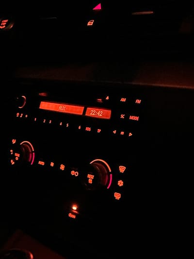 The image depicts the interior of a car at night. The dashboard is illuminated by red and orange lights. A car stereo system is visible, displaying "AUX" and the time "22:42". There are various buttons and controls for the stereo and climate control, including knobs and symbols for fan speed, temperature, and air direction. A button labeled "REST" is also visible. The overall environment is dark, consistent with nighttime or a dimly lit car interior. The location in the city of Bazan, Bulgaria, is not discernible from the image itself. There are no people, actions, or interactions visible in the photograph.