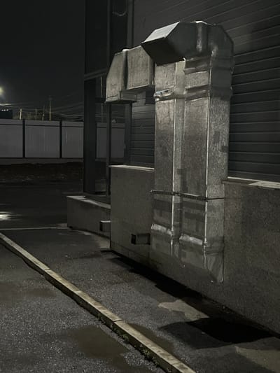 The image shows a close-up of a large, rectangular metal ventilation duct system mounted outdoors. The ductwork appears to be made of galvanized steel and is secured with metal straps and possibly tape. The ducting has several bends and turns, suggesting a complex system. It is installed under a dark, ribbed ceiling structure.

The scene appears to be an industrial or commercial exterior. The ground below the ducting is a dark, textured surface, possibly asphalt or concrete, which appears wet, reflecting some faint light. A concrete curb or raised edge runs diagonally across the lower right portion of the image.

The lighting suggests it is nighttime or very dim conditions, with a few distant light sources visible as small white dots in the background on the left. The overall mood is utilitarian and somewhat stark due to the industrial materials and the low light. There are no people visible in the frame. No visible text is discernable on the ductwork or the surroundings. The location context of Qoyankus, Kazakhstan, places this scene in an outdoor environment within that region.