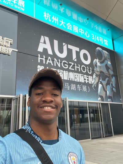 The image shows a young Black man standing in front of a building in Hangzhou, China. He is smiling and looking directly at the camera. He is wearing a light blue soccer jersey with a Manchester City logo on the left side and a black strap across his chest. The man is standing in front of a glass door, and the building is decorated with a large banner that reads "AUTO HANGZHOU".  The banner features a large white robot wearing an astronaut helmet, and Chinese characters. The banner also reads "Auto Show" in English. The location is likely the Hangzhou International Expo Center at the coordinates provided.