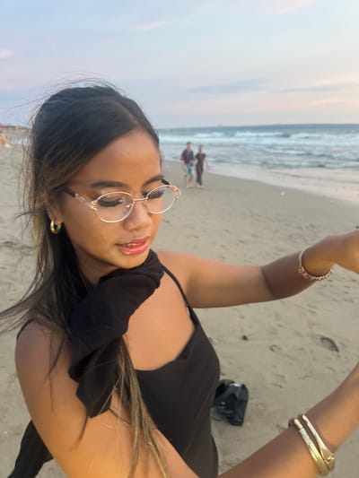 A woman with long, dark hair, wearing light-rimmed eyeglasses, a black sleeveless top, and a black fabric around her neck, is featured prominently in the foreground. She has a gold hoop earring on her left ear and multiple gold-toned bracelets on her right wrist, one featuring red accents. Her lips are glossy. She is looking downwards and to her right, with her right arm extended, partially out of frame. The scene is a sandy beach in Kerobokan Kelod, Indonesia, at dusk, characterized by a light blue and faint pink sky. In the blurred mid-ground, two individuals, one male in a plaid shirt and shorts, and one female in dark clothing, walk along the shoreline where ocean waves are visible. A dark, indistinct object rests on the sand behind the main subject's right arm.