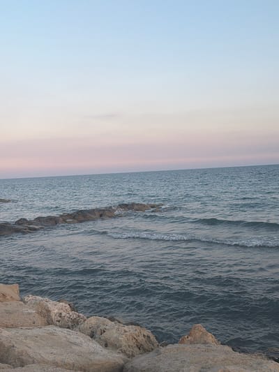 The image depicts a tranquil seascape with a rocky shoreline in the foreground, likely located in Pervolia, Cyprus. The sky displays a gradient of soft blues and pinks, suggesting either sunrise or sunset. The calm sea is a deep blue-gray, with gentle waves lapping against a submerged line of dark rocks extending into the water. The foreground consists of large, light-colored rocks and boulders, characteristic of coastal areas. No people or specific landmarks are visible, presenting a natural, undisturbed coastal environment.