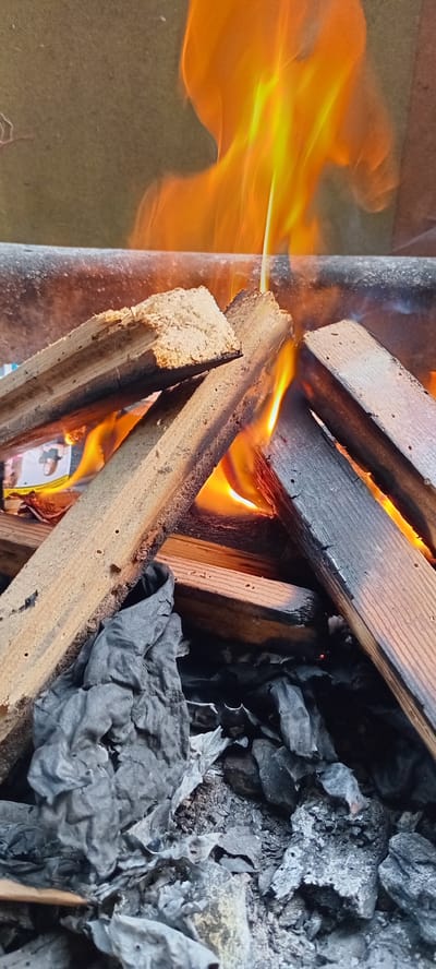 The media file captures a close-up view of a fire burning wood and paper. The main subjects are several pieces of wood, some of which are charred and actively burning with vibrant orange and yellow flames licking their edges. Interspersed with the wood are crumpled pieces of dark gray paper, also showing signs of being consumed by the fire, turning into ash in some areas.
The scene is set within what appears to be an outdoor environment, possibly a fire pit or a brazier, given the presence of ashes and embers. The background shows a textured surface, possibly fabric or a rough ground covering. The lighting is dominated by the intense glow of the fire, creating strong contrasts and illuminating the subjects.
The activity taking place is the combustion of fuel, with the wood and paper being the objects consumed by the flames. The fire is intense, with visible tendrils of flame extending outwards. The overall impression is one of heat and destruction. There are no people visible in the frame.
Notable details include the texture of the burnt wood, showing grain and splintered edges, and the delicate, crinkled texture of the burning paper. The presence of ash suggests that the fire has been burning for some time, or is in the process of completing its cycle. There is no indication of the time of day or weather conditions, other than the fire itself creating its own light. There are no discernible emotions conveyed due to the absence of human subjects. No visible text is present in the image. The location context is Iskrets, Bulgaria, but this information is external to the visual content of the image itself.