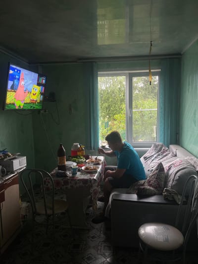 In a dimly lit room, a young person wearing a blue t-shirt and dark shorts sits at a table, eating. The table is covered with a floral-patterned tablecloth and laden with food, including plates of pasta, salad, and bottles of drink. To the left, a television displays cartoon characters, Patrick Star and SpongeBob SquarePants. Behind the person, a window offers a view of lush green trees. The room is furnished with a sofa, chairs, and a cabinet. The walls are covered in green tiles, and the ceiling appears to be made of reflective panels. The floor is tiled with a geometric pattern. The overall scene depicts a casual meal in a domestic setting, likely a kitchen or dining area, within the city of Krasnoye, Russia.