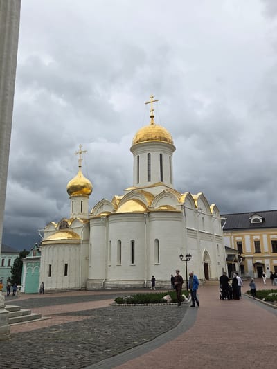 The image shows the Trinity Lavra of Saint Sergius in Sergiev Posad, Russia, under a cloudy sky. A large white church with multiple golden domes and crosses dominates the center of the frame. To the right of the church, a yellow building with a dark roof is visible. In the foreground, a paved courtyard with cobblestone and brick patterns is present. Several individuals are scattered throughout the scene, walking or standing. One group of people, including a person pushing a stroller, is walking towards the right. Another person in a blue jacket and another in a brown suit and hat are walking on the left side of the paved area. The overall atmosphere appears to be a typical day at a historical and religious site.