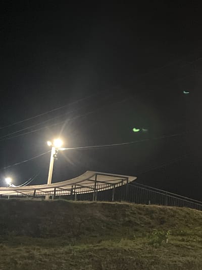 The image shows a nighttime view of an outdoor structure, possibly a covered seating area or pavilion, situated on a grassy incline. A metal railing lines a sloped pathway leading up to the structure. A lamppost with a bright light is prominently visible on the left side of the image, illuminating the immediate surroundings. Several thin wires crisscross the dark sky above. In the upper right portion of the sky, there are three faint, greenish, elongated shapes, possibly distant lights or atmospheric phenomena. The environment appears to be an open area with grass and a subtle slope, suggesting it could be a park, recreational area, or roadside attraction. The specific location is identified as Golubitskaya, Russia. There are no people visible in the image, and the focus is on the static elements of the scene.