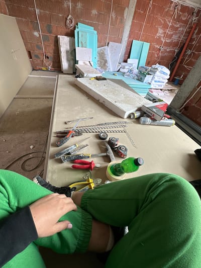 The image shows a work area with various tools and materials. On a light-colored surface, there are tools such as drills, measuring tapes, a hammer, pliers, utility knives, and screwdrivers. There are also cans of construction adhesive and a green water bottle. To the left, there are stacks of plasterboard and bags of plastering compound. The background reveals exposed brick walls with visible wiring and piping. In the foreground, a person wearing green sweatpants and black shoes is visible, with their hand resting on their leg. The location is likely a construction site or a room undergoing renovation. There is no indication of the location being Aprilovo, Bulgaria.