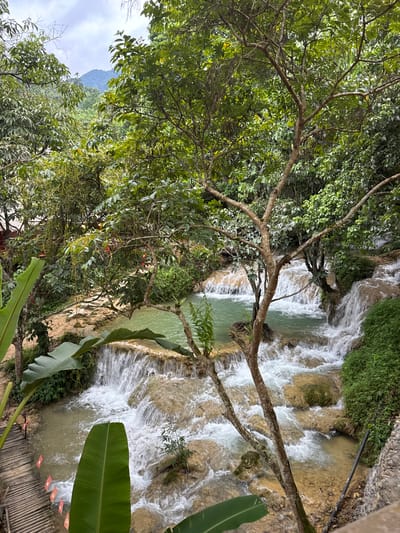 The image depicts a natural landscape featuring a multi-tiered waterfall with clear, turquoise water flowing over light brown rocks and cascading into pools. Lush green foliage, including large banana leaves and dense trees with visible branches and smaller leaves, surrounds the waterfall. A wooden walkway, made of planks and elevated slightly, is visible on the left side of the image, adorned with small red flags. In the background, a distant mountain covered in vegetation can be seen under a cloudy sky. The scene is evocative of a tropical or subtropical environment, likely a natural park or scenic area. The presence of the walkway suggests it is an accessible tourist destination within the Cổ Lũng Commune, Vietnam, likely in a rural or mountainous region, as indicated by the natural terrain and vegetation.