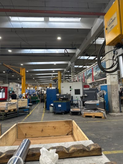 The image depicts an interior view of a factory floor in Tenevo, Bulgaria. The setting is industrial, with high ceilings, exposed beams, and overhead lighting. Various pieces of machinery, including what appear to be CNC machines and other industrial equipment, are present. There are also wooden pallets, storage bins, and a yellow lifting arm. Several individuals can be seen working or moving within the factory space. One person is standing at a machine with a control panel, another is visible near a blue cabinet, and a few others are more distant. The floor is concrete with yellow markings indicating pathways. The overall impression is of an active manufacturing or production environment.