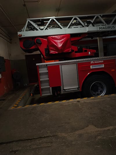 The image shows a red Magirus fire truck with an extendable ladder parked in a garage in Tukums, Latvia. The fire truck is positioned over a recessed pit with yellow and black striped safety markings on its edge. The garage appears to be dimly lit, with exposed ceiling pipes and wiring. To the left of the fire truck, a portion of an orange wall is visible, along with some equipment mounted on it. In the background, a dark wooden door and a red cylindrical object on a stand can be seen. The fire truck itself is clean and appears to be in good condition, with the Magirus logo visible on its side. There are no people or animals visible in the image.