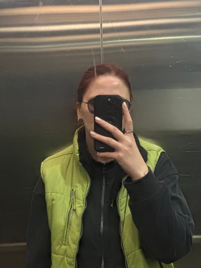 A person is taking a selfie in what appears to be an elevator with brushed metal walls. The person is wearing a black hoodie and a lime green vest. They are holding a black smartphone with a triple-lens camera to obscure their face. The person has light skin and reddish-brown hair and is wearing sunglasses. The background is the blurred reflection in the elevator's metallic walls. There is no indication of the location being Overyata, Russia.
