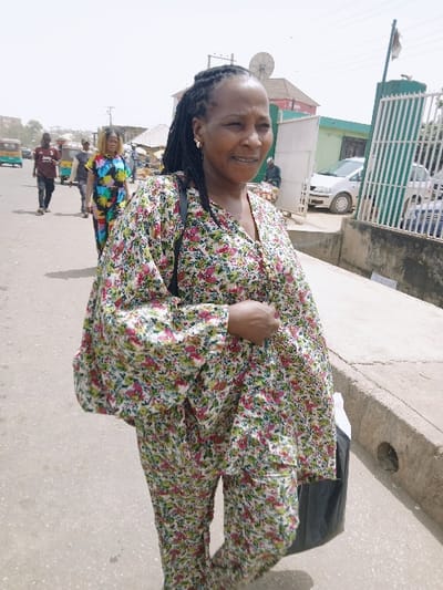This image captures a daytime street scene in Kadoma, Zimbabwe. The central figure is a woman of apparent African ethnicity, positioned slightly right of center, with her head turned slightly to her right, displaying a faint smile. She has dark braided hair and wears small gold-colored hoop earrings. Her attire consists of a two-piece ensemble (blouse and trousers) with a prominent floral pattern in shades of green, pink, yellow, and white on a light background. A dark strap from a bag or purse is visible over her left shoulder, and her right hand holds a black plastic bag.

She stands on a paved road, which recedes into the background. To her immediate right, a concrete curb is present, featuring a circular drainage opening. Beyond this curb, a green metal gate or fence partially obscures a light green building, behind which a white sedan is visible. Further in the background, a large satellite dish is mounted on a structure, and additional indistinct buildings line the street.

In the mid-ground, on the paved road, several other individuals are visible. One man, dressed in a dark red shirt and dark trousers, walks away from the camera. Another woman, wearing a brightly colored, multi-patterned dress, walks towards the camera. A green and yellow auto-rickshaw is also visible further down the street. The sky overhead appears bright and hazy. The scene depicts an active urban environment with pedestrian traffic and permanent structures.